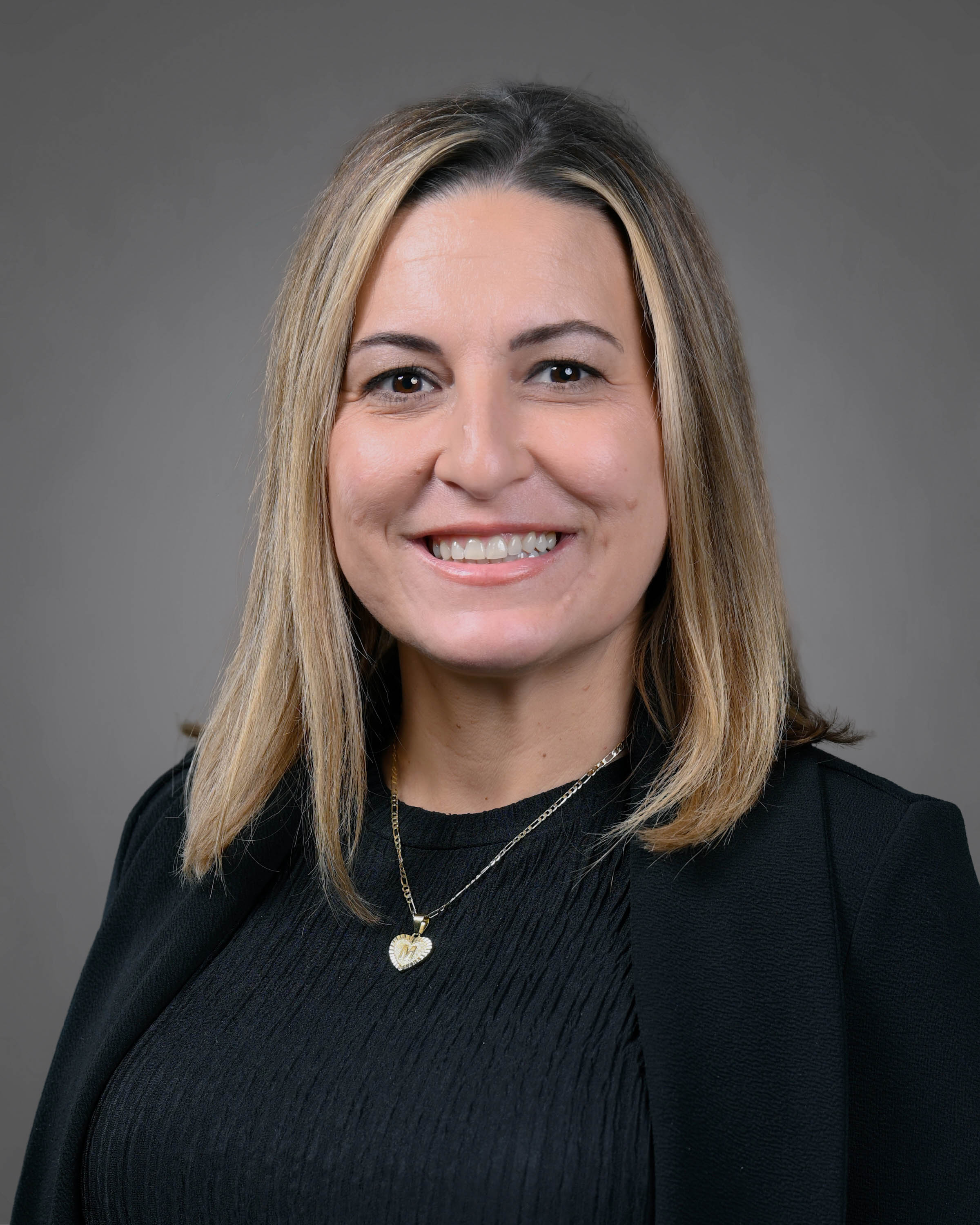 Professional headshot of a smiling woman with shoulder-length blonde hair, wearing a black blazer, black blouse, and heart-shaped pendant necklace against a gray studio background.
